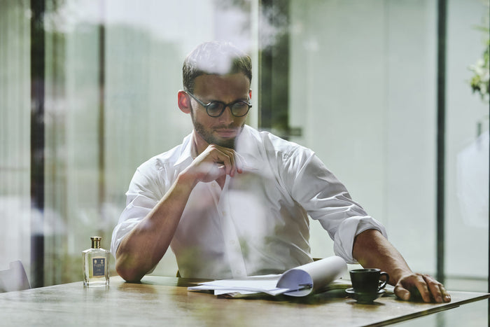 A man sat at a tale in a white shirt and black rimmed glasses, looking at work with a bottle of no. 89 next to him.